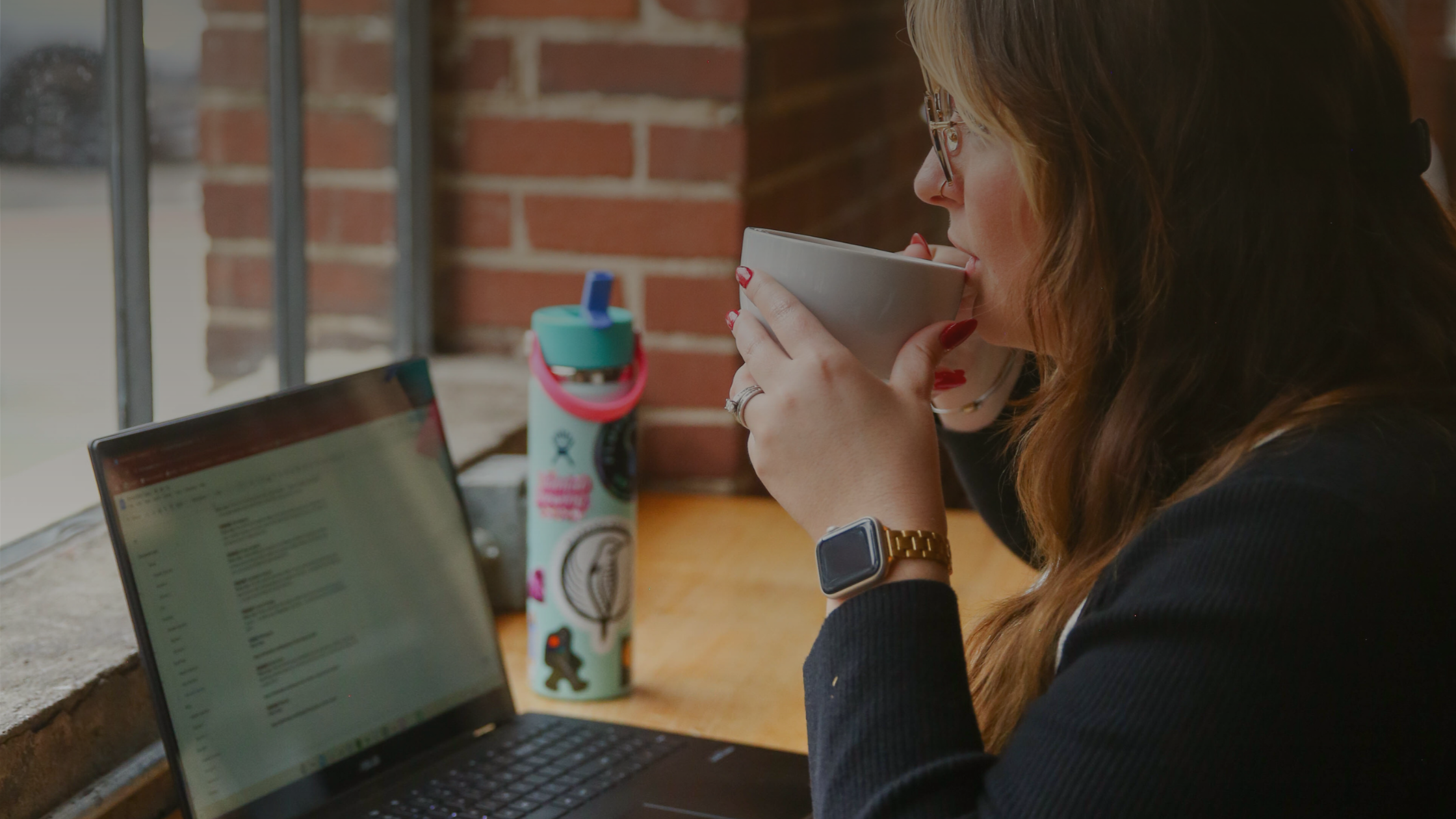 laptop sitting on a table, woman with dark hair sipping a coffee sitting in front of it.