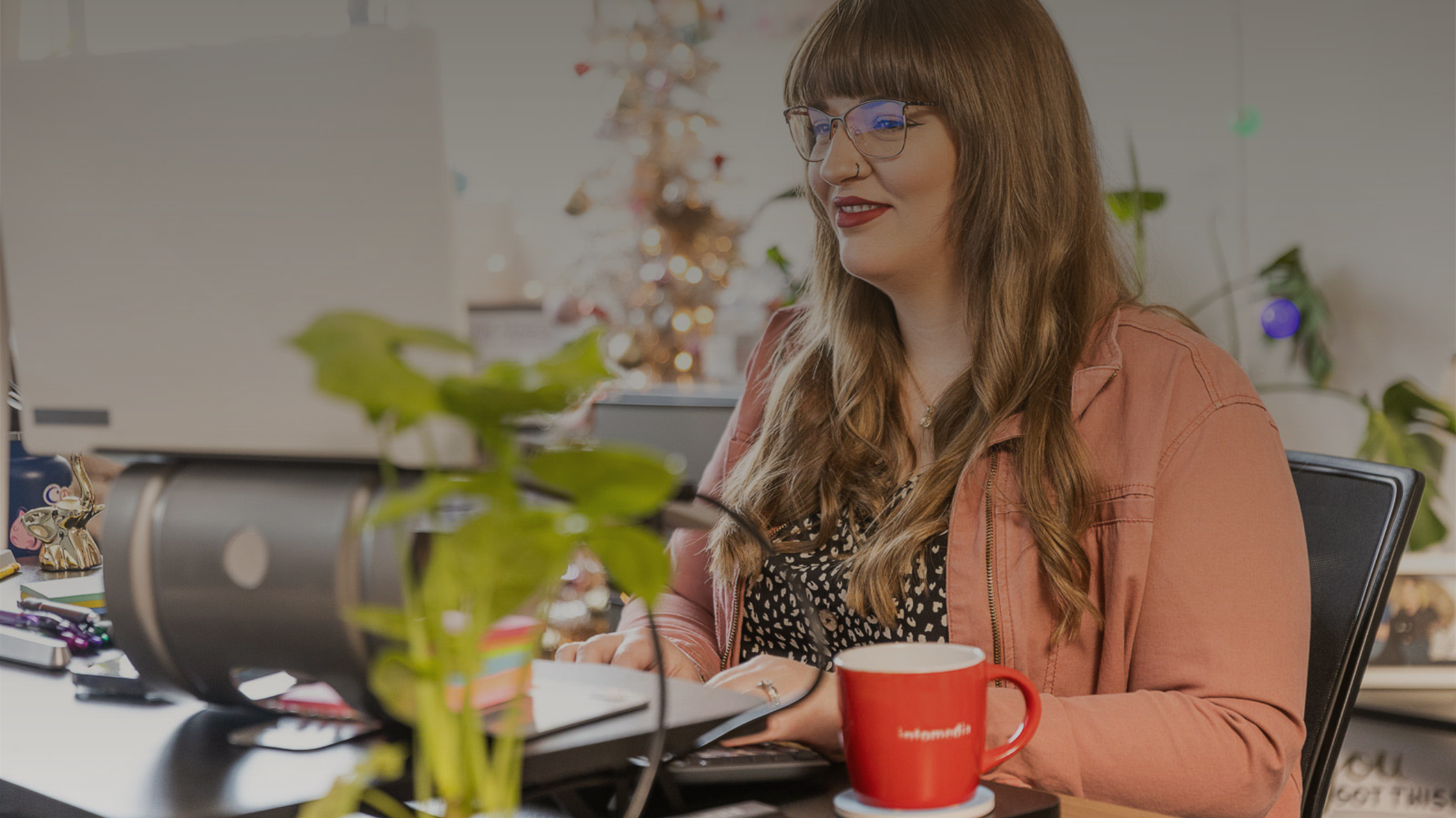 Woman with dark hair and a pink jacket typing on a keyboard and looking at a computer monitor off screen.